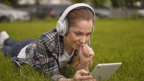 The girl listens to music in large headphones on nature and scrolls the news on the tablet. woman is very surprised and glad from what she saw 4K