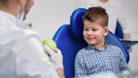 Male dentist in uniform giving fresh green apple to happy child. Smiling baby boy with healthy teeth posing in stomatologist armchair at clinic. Medium shot on RED camera