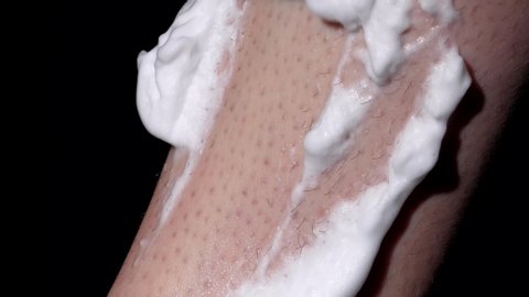 This close up, macro video shows an anonymous hand applying shaving cream and then shaving a hairy leg with a razor against a black background.