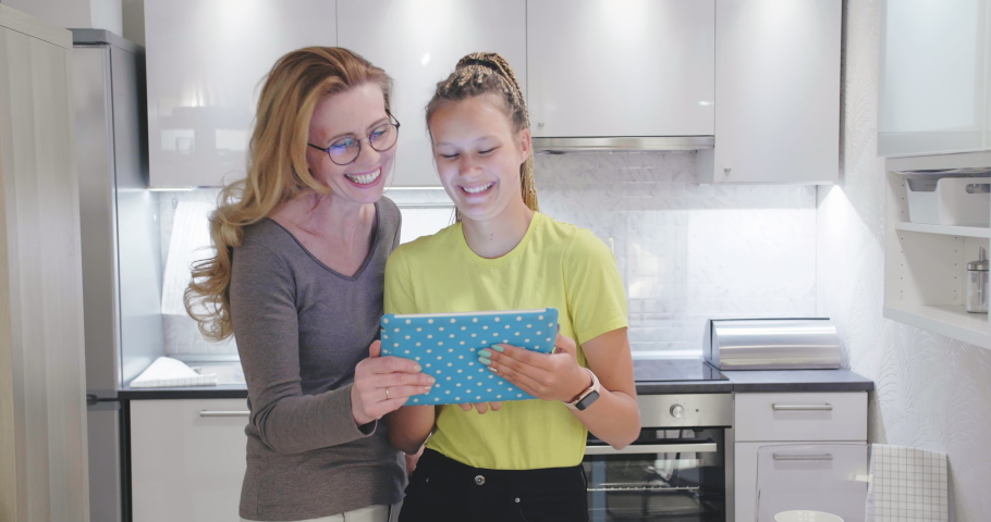 Middle aged mother and her adult daughter watching funny videos using tablet PC at their modern kitchen