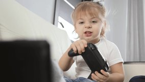 A blonde girl is playing a game console and smiling. A child sitting on a white sofa at home with a joystick in his hands.