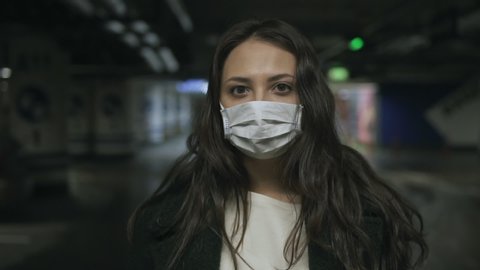Portrait of attractive young woman in protective medical mask, standing alone in empty underground Parking lot during quarantine due to covid-19 coronavirus pandemic, indoors closeup slow motion