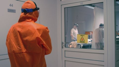 Doctor in an Orange Protective Suit Enters Isolation Room with Coronavirus Patients - Handheld Medium Tracking Shot in Slow Motion