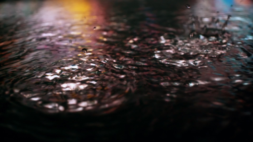 Rain drops falling into puddle on asphalt at night. Big city street lights mirror reflection on water surface. Close-up slow motion of raining outside in wet season