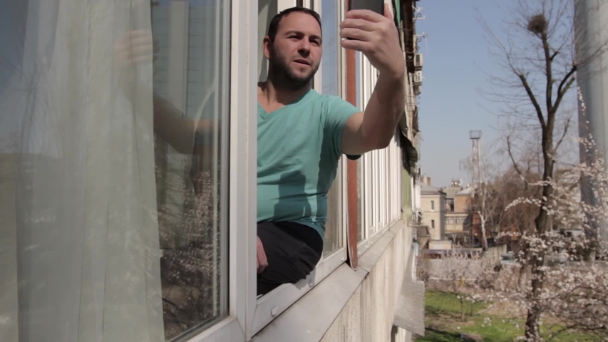 A man sits on a window frame and communicates via smartphone, finishes the conversation, says goodbye and goes to the apartment. camera movement through the window frame, from right to left. 