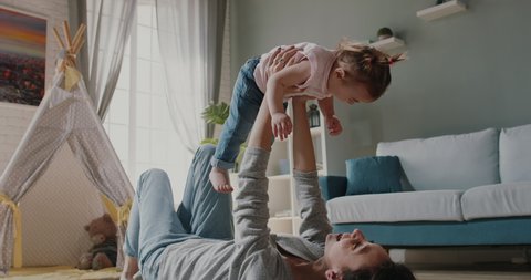 Active caucasian young mom and her little cute daughter playing at home. Joyful babysitter working at home, holding the baby up - happy family, activity together concept 4k footage