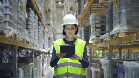 Young asian woman using tablet while walking in warehouse of modern factory. Front view of female employee doing work in storehouse, holding digital device in hand and looking at screen. 
