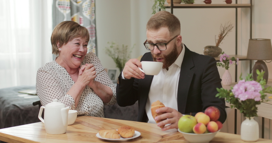 Handsome man in glases and suit eatting and drinking while sitting at table of mothers home. Cheerful retired woman in 70s looking happy and laughing while spending time with son