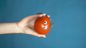Red tomato with eyes in a woman hand close up. Tomato looks around on a blue background. Vertical video.