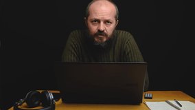 Studio shot of bearded caucasian man in sweater sitting behind desk with headphones and talking isolated against black background, video blog concept