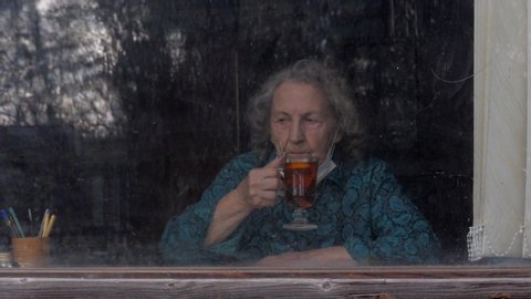an elderly woman lowering her medical mask drinks a beautiful tea and looks out the window. coronavirus pandemic, covid-19