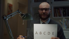Bearded 30s man in glasses showing sheet of paper with alphabet letters at camera at home. Online education process. Webcam view of teacher talking. 