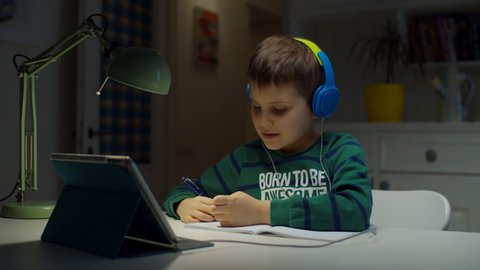 Schooler boy in color headphones using tablet computer for online education at home. Kid enjoys making school homework online, hand writing in textbook. 