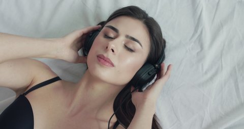 Top view of nude girl in bra swaying to her music in headphones with closed eyes, then puts off them and rejoices into camera on bed. Slowly in 4K