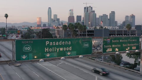 Los Angeles. 2019. Aerial. Drone rises up over the freeway, from the green road sign with Pasadena and Hollywood written on it, to the view of downtown with its skyscrapers and many intersections. 4K