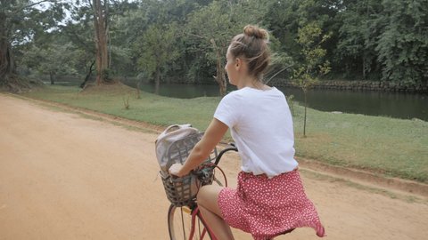 Young woman cycling on dirt road in Sri Lanka traveling and exploring nature and sightseeing. Girl on bicycle going to visit Sigiriya area, waving hello to camera and partner 