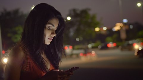 An attractive/ beautiful girl standing outside/ outdoors using mobile phone/ smartphone in the evening. A modern corporate/ office woman/ female typing on a cellphone against the moving traffic.