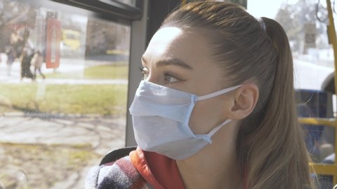 A young Woman in a protective mask rides public transport and looks out the window. Portrait of a woman in a protective mask on a social bus. Pandemic COVID-19. Social distance.