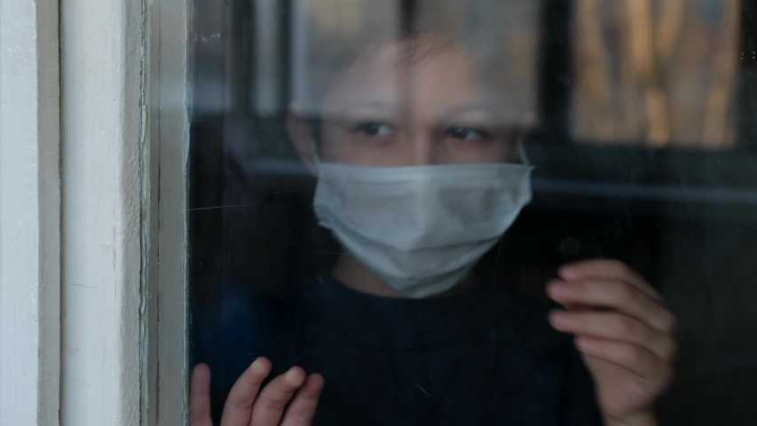 Young boy in a medical mask looks out the window. Self-isolation in quarantine, coronavirus, covid 19. Royalty-Free Stock Footage #1049810173