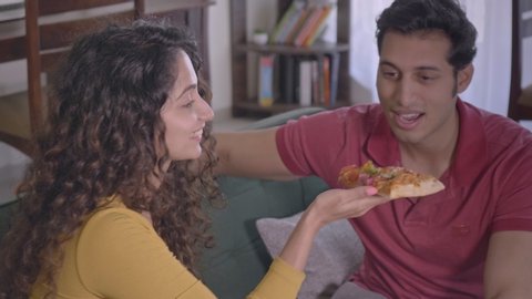 A happy young woman girl female teasing her man male partner by showing a piece slice of pizza. A joyous playful married husband and wife laughing enjoying in interior house. relationship goals.