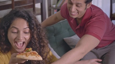 A happy young woman girl female teasing her man male partner by showing a piece slice of pizza. A joyous playful married husband and wife laughing enjoying in interior house. relationship goals.