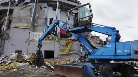 OMSK/RUSSIA - MARCH 02 2020: Modern blue grader disassembles debris at demolition site of destroyed hockey stadium under cloudy sky on March 02 in Omsk