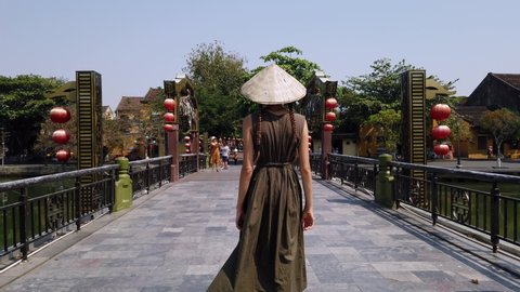Hoi An / Vietnam - MARCH 09 2020: Beautiful young woman tourist in long green dress and Vietnamese hat walks on bridge in Hoi An old town, look at river, Vietnam. Сyclists drive across bridge. Tourism
