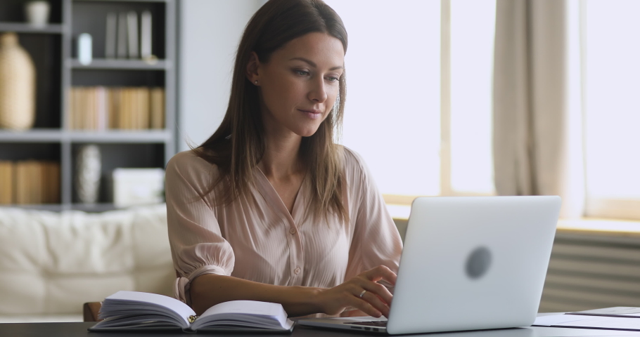 Pleasant young freelance businesswoman working from home or studying remotely on online courses on computer, communicating with clients, writing making appointment in personal paper organizer. Royalty-Free Stock Footage #1049816893