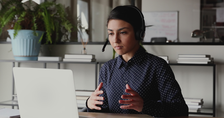Confident indian female web coach, online teacher or customer support service operator wearing headset speaking to webcam looking at laptop. Ethnic business woman making conference video call concept. Royalty-Free Stock Footage #1049816968