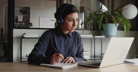 Female indian distant worker wears headphones video calling makes notes, elearning with online teacher, working from home office attending remote conference meeting. Videoconference training concept.