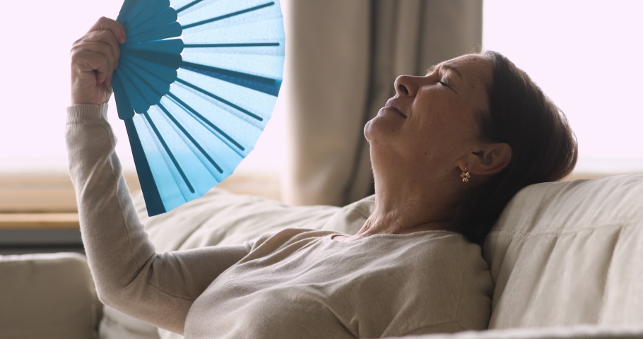 Side view head shot overheated middle aged mature woman using paper fan, suffering from high temperature indoors. Stressed senior older grandmother feeling hot without air conditioning system at home. Royalty-Free Stock Footage #1049817070