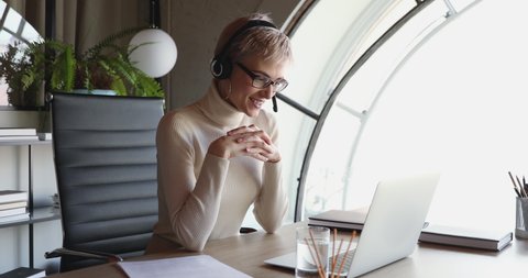 Young business woman wearing headset conferencing in distance remote web chat sitting at office desk. Female worker, coach, manager participating webcam video call speaking looking at laptop.