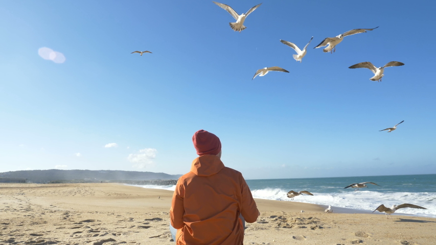 young guy in bright orange jacket feeds hungry seagulls on sand beach against crystal blue ocean with waves backside view