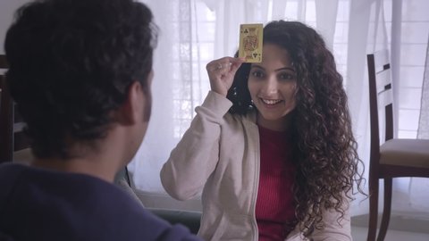 A happy smiling attractive young married couple playing enjoying cards together indoors home or house interior setup. A girl sticking card on her forehead while playing Blind man's bluff, Indian poker