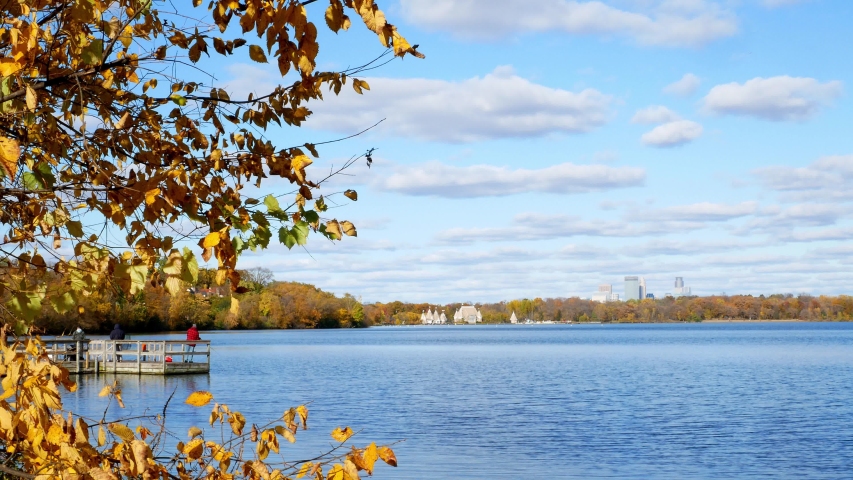 Beautiful Lake Harriet in Minneapolis, Minnesota on a sunny autumn afternoon with colorful leaf foliage, blue sky and fluffy white clouds, a fishing dock and downtown buildings in the distance.