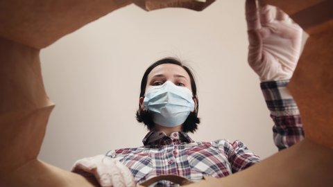 Young woman carefully opens box delivery from online store. Coronavirus pandemic quarantine, girl portrait with personal protective equipment gloves and a medical mask on face, second wave covid