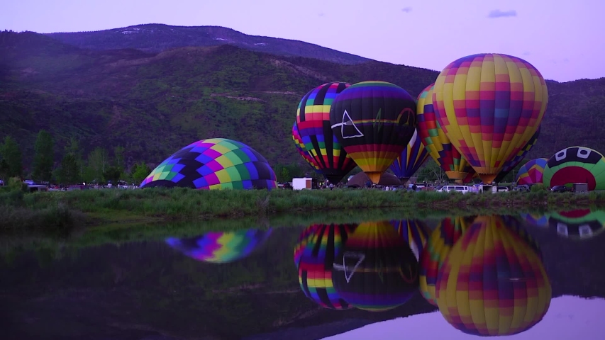 Hot air balloons inflate and glow at dusk, reflected in a park lake.  A balloon starts to inflate at left.  Basalt Mountain in the background.  Balloon glow randomly.