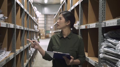 elegant asian female warehouse worker with clipboard doing stock taking job and writing report. young woman employee holding touch pad walking out and meet colleague asking problem in stockroom.