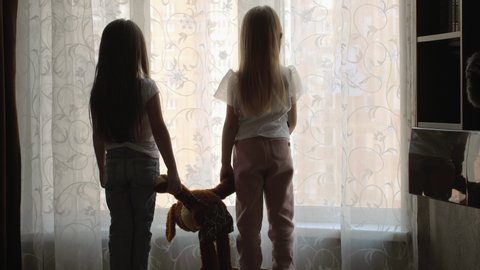 Two little girls with a toy in their hands stand by the window and look into the distance. Home mode.