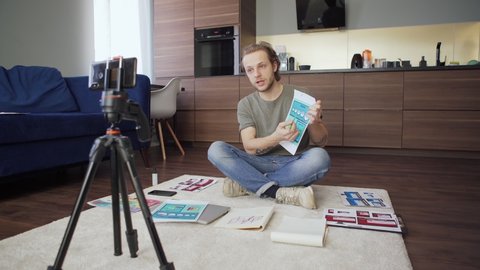Panning shot of young male designer sitting cross-legged on floor at home, having video conference with colleagues on smartphone and showing printed templates