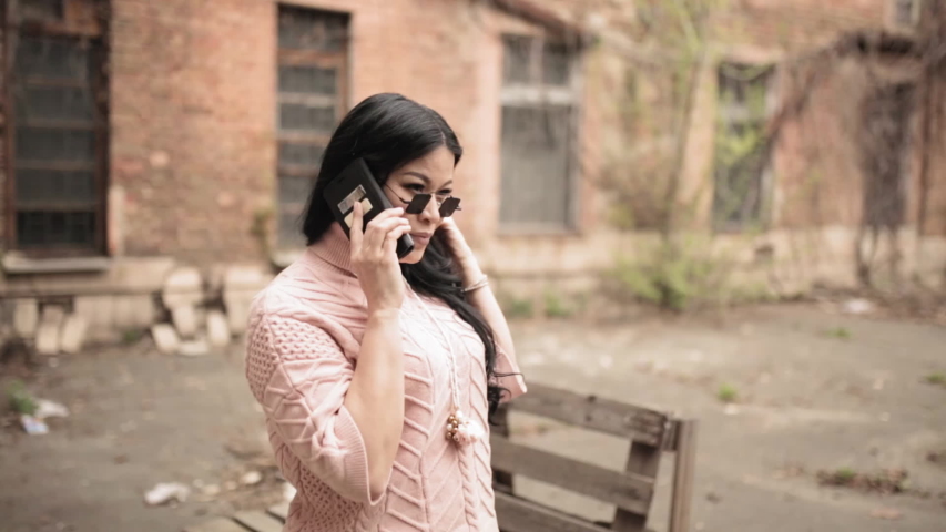 Beautiful middle-aged Asian woman in a pink light sweater with dark stripes, and fashion square sunglasses talking on the phone against the backdrop of ruins