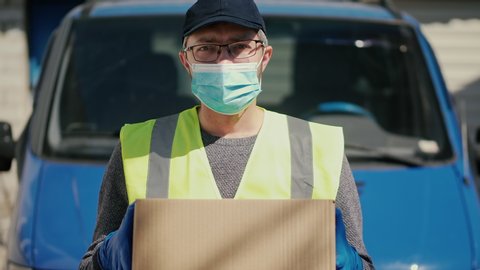 Portrait of a courier in a medical mask and protective gloves holding a parcel, a blue car in the background.