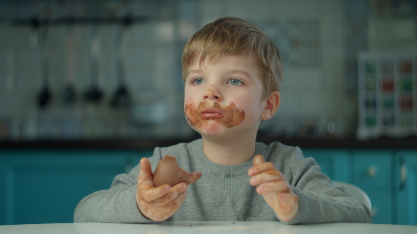 Blonde boy eating Easter chocolate bunnies with dirty face at home on blue kitchen. Happy kid celebrating Easter with colored eggs and chocolate bunnies.  | Shutterstock HD Video #1049873791