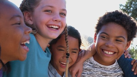 Camera tracks along faces of group of children hanging out with friends outdoors with arms around each other against flaring sun - shot in slow motion