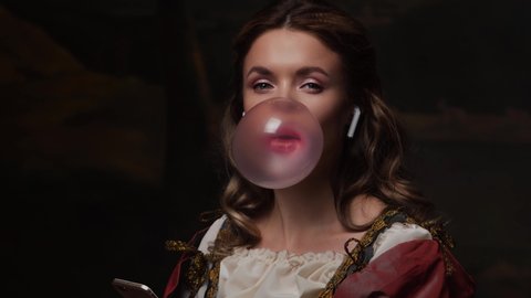 Medieval teen, young girl in vintage dress uses smartphone and blows bubble gum. Beautiful young woman in the style of a Renaissance painting.