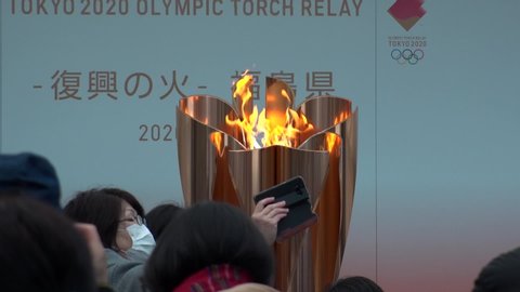 FUKUSHIMA, JAPAN - 24 MAR 2020 : Olympic Flame displayed at Fukushima station. Tokyo Olympic 2020 have been postponed to 2021 due to coronavirus. Crowd of people wearing surgical masks. Slow motion.