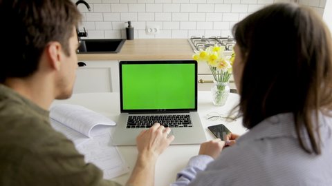 Young casual couple using Macbook laptop with green screen and sitting at home kitchen, choosing best sale offers, purchasing goods in online shop together, searching cheap travel tours
