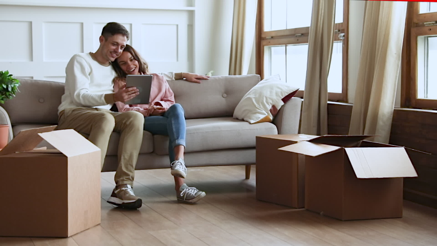 Happy young married couple sitting on couch, laughing at funny video on digital tablet while small children siblings running playing with huge cardboard boxes in renovated new living room, moving day. Royalty-Free Stock Footage #1049912191