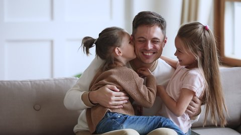 Happy young handsome father sitting on sofa, holding on lap small children daughters, enjoying tender sweet moment at home. Cute little kids girls embracing kissing smiling father, slow motion.