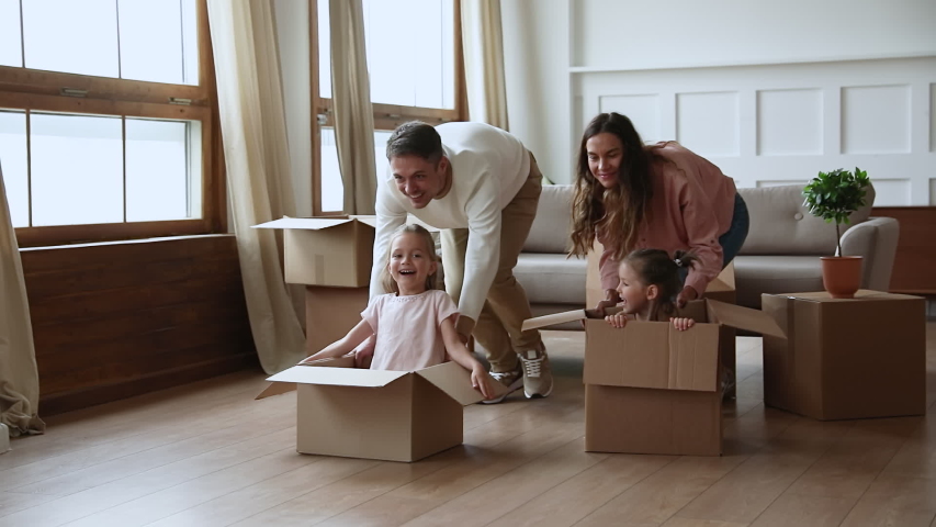 Slow motion active carefree young couple parents pushing huge cardboard boxes with kids, running forward, slow motion. Happy father and mother playing, having fun with adorable children daughters. Royalty-Free Stock Footage #1049912203